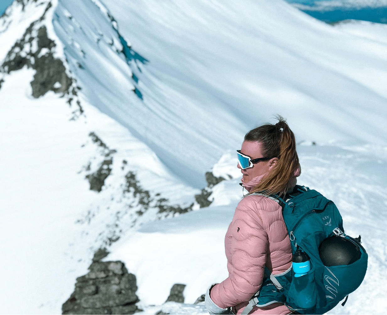 Une guide de trek équipée, en tenue d'hiver observant l'horizon en haut d'une montagne enneigée.