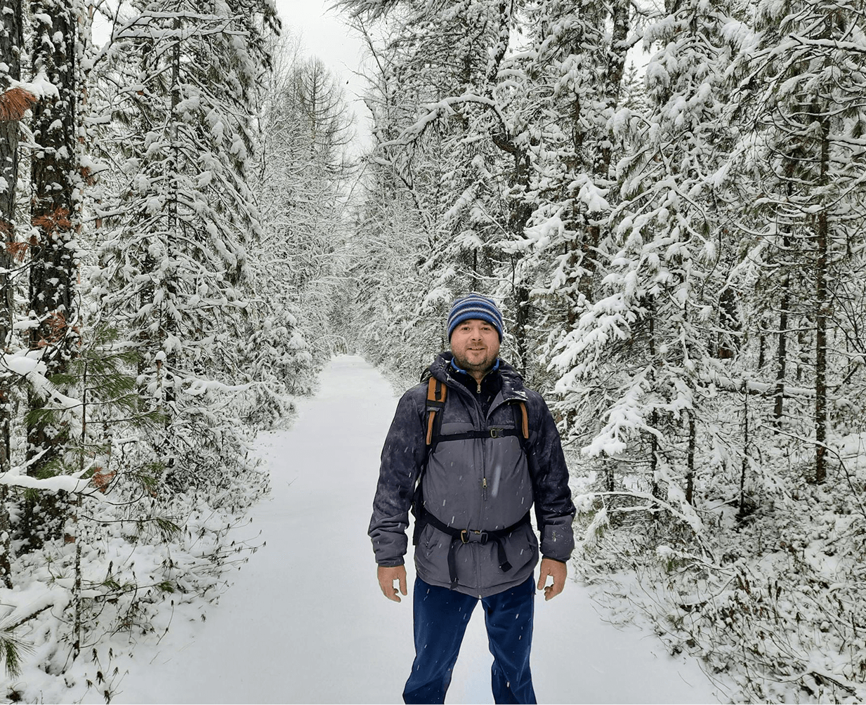 Un homme souriant, habillé en tenue d'hiver posant pour une photo au sein d'une forêt de sapins enneigée.