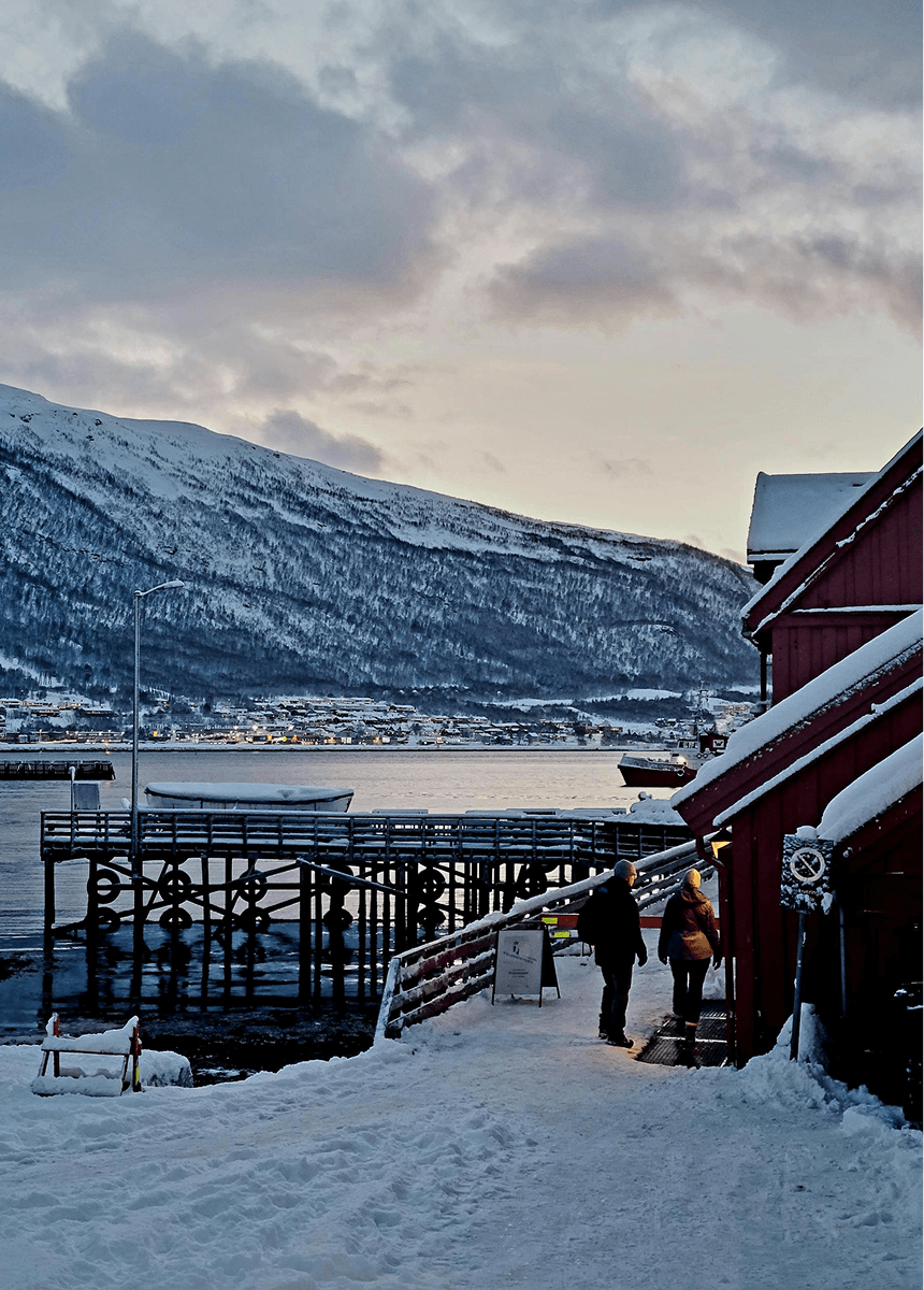 Une petite maison typique de la Norvège près d'un port aux îles lofoten par temps de neige. Un couple semble se promener près du port.